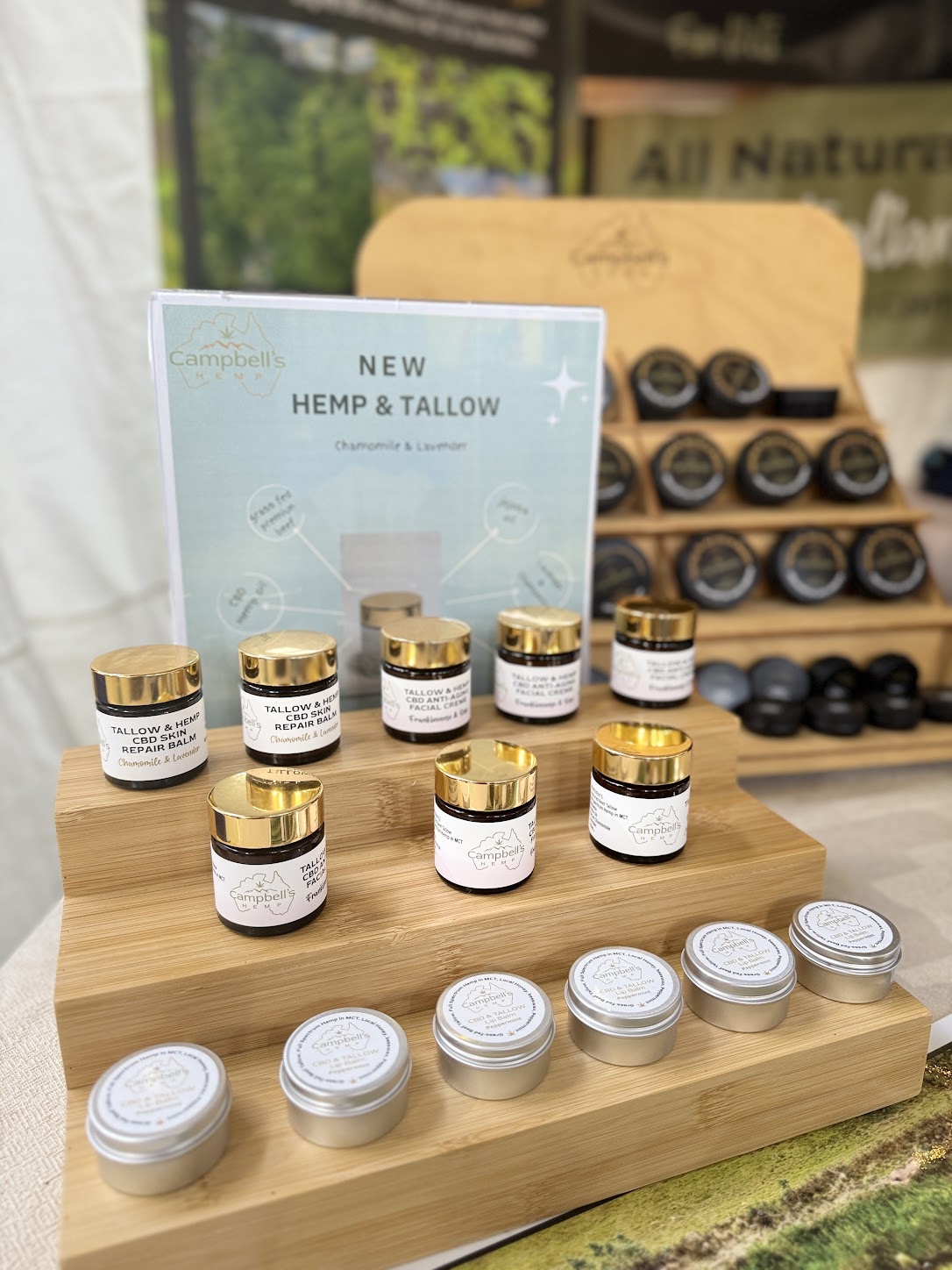 Display of small jars with gold and silver lids on a wooden stand, with a sign indicating 'New Hemp & Tallow' in the background.